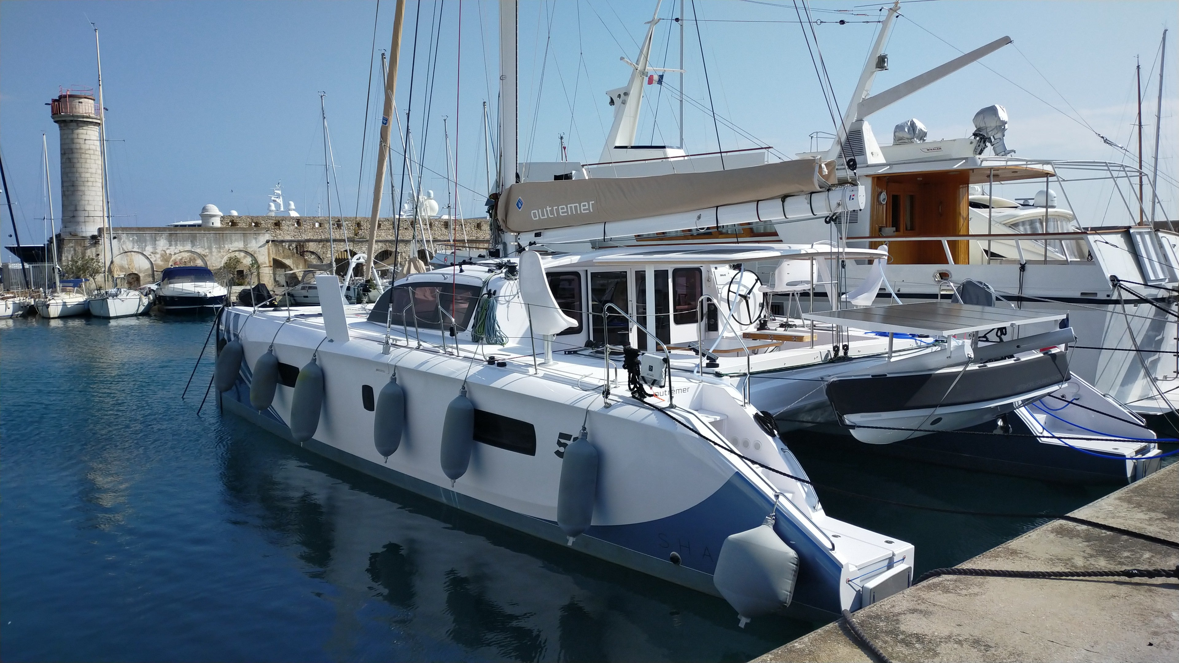 White catamaran docked at a marina with clear blue water and sky. Textile grey fenders protect the vessel as boat bumpers.