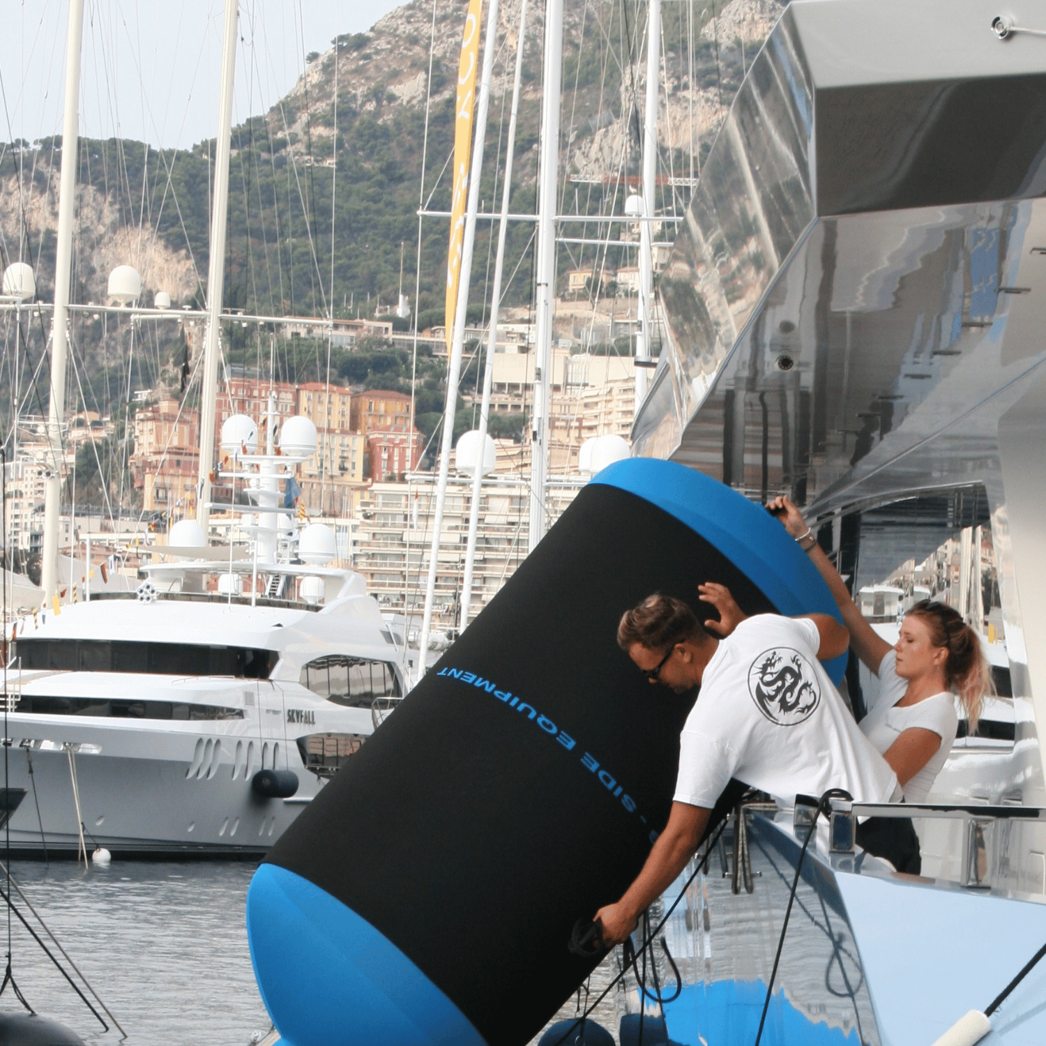 Two people interacting with a large blue boat fender and black fender cover on a yacht with yachts in the background.